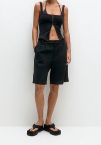 Kobieta w czarnym bezrękawniku, czarnych szortach do kolan i czarnych sandałach na koturnie, stojąca z rękami w kieszeniach.