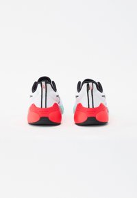 Hvite sneakers med røde og svarte detaljer. Teksturert materiale, gummisåle og mesh-overdel. Har svart stripe og "NANO GYM" merke.