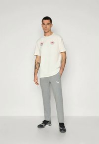 Camiseta de color beige claro con detalles de logotipo, combinada con pantalones de chándal grises que tienen un logotipo blanco. El modelo lleva zapatillas deportivas negras.