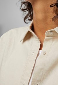 Gros plan d'une personne portant une chemise beige à col ouverte sur un haut blanc, montrant le cou et des mèches de cheveux bouclés et foncés.