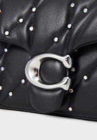 Svart quiltad läderhandväska med silvernitar och ett stort silverfärgat C-format lås mot en vit bakgrund.