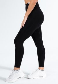 Zwarte legging van rekbare stof, hoog in de taille, met een gladde textuur en een contrasterend wit logo op de tailleband. Witte sneakers met zwarte accenten.