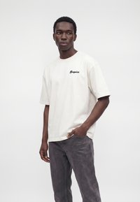 Junger Mann trägt ein lockeres weißes T-Shirt mit kleinem schwarzem Text auf der Brust und dunkelgraue Jeans, steht mit einer Hand in der Tasche.