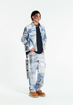 Joven con rastas que lleva una chaqueta y pantalones de estampado azul y blanco, camisa azul marino, botas color cuero claro y un accesorio de correa larga colgante.