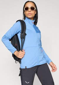 Ljusblå zip-jacka med texturerad yta, har svarta detaljer, blixtlåsfickor och en logotyp. Matchas med mörka byxor och en svart ryggsäck.