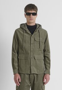 Hombre vestido con una chaqueta de corduroy verde oliva con capucha y pantalones a juego, acompañado de gafas de sol rectangulares negras, de pie frente a un fondo blanco.