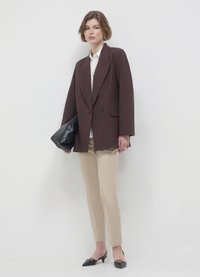 Veste croisée marron, blouse blanche, pantalon beige et escarpins noirs à bouts pointus. Tenant une pochette en cuir noir, sur fond blanc.