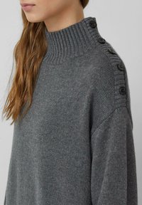 Grauer Strickpullover mit einem gerippten hohen Kragen und Knopfakzenten entlang der Schulter. Weiche Textur mit lockerer Passform und minimalistischem Design.