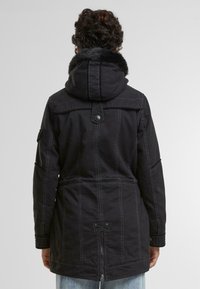 Svart jakke med fleecefôret hette, med kontrasterende hvite sømmer, trykknappslukking og midtlang design med sidelommer.