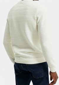 Vit strukturerad tröja med horisontella ribbmönster och en figurnära nederkant, bärs tillsammans med mörkblå jeans. Bakomifrån visas plaggets design detaljer.