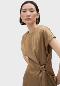 Abito in satin marrone con maniche corte, caratterizzato da un dettaglio a nodo attorcigliato sul lato, texture liscia e silhouette rilassata.
