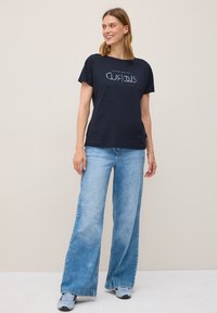 En leende kvinna står mot en ljus bakgrund, iförd en marinblå t-shirt med ordet "CURIOUS" och vida jeans.
