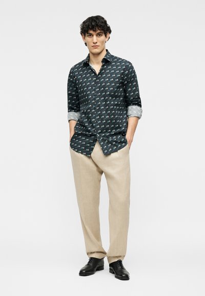 Νεαρός άνδρας που στέκεται με τα χέρια στις τσέπες, φορώντας σκούρο πουκάμισο με μοτίβο και μακριά μανίκια, μπεζ παντελόνι και μαύρα επίσημα παπούτσια, μπροστά σε λευκό φόντο.