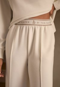 Crème-kleurige broek van ribstof met een elastische tailleband en een subtiele logo-opdruk in grijs. Ontspannen pasvorm met plooien.
