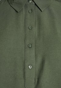 Zielona oliwkowa koszulka polo z kołnierzykiem, z czterema dopasowanymi guzikami wzdłuż zapięcia, ukazująca teksturę materiału i szczegóły szwów.