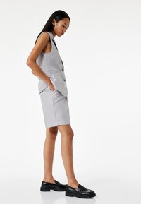 Chaleco sin mangas en gris claro con bolsillos, combinado con un pantalón corto a juego. Se llevan mocasines negros con una superficie texturizada.