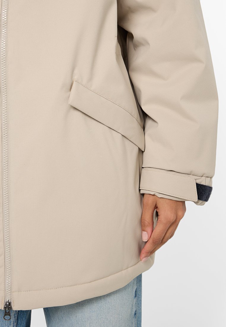 Beige lange mouw jacket met een rits en zijzak; heeft een gestructureerde stof en een manchet met een donkerblauw accent eronder.