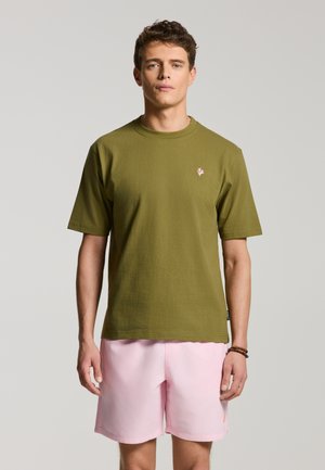 Young man standing still wearing olive green short-sleeve shirt and light pink shorts against plain gray background.