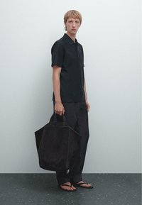 Polo nero e pantaloni scuri, abbinati a una grande borsa a mano in camoscio scuro con manici robusti. Sandali casual completano il look.