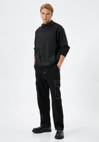 Svart tröja med hög krage och stängning med två knappar, kombinerad med svarta cargo-byxor med kontrasterande vit sömnad och fickor.