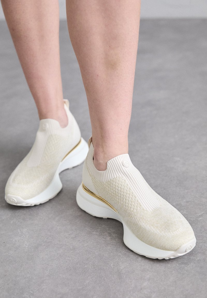 Lys beige sportsko med en ribbet strikket overdel, teksturdetaljer, og en hvit såle med gull aksent. Slip-on design.