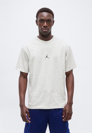 Mladi moški v svetlo sivi majici Jordan in modrih športnih hlačah, stoji pred enobarvnim belim ozadjem.