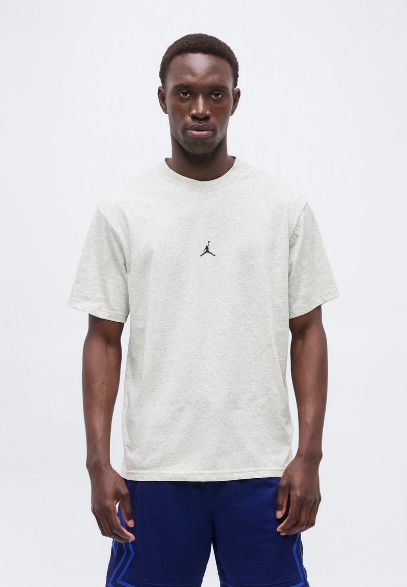 Νέος άνδρας με ανοιχτό γκρι μπλουζάκι Jordan και μπλε αθλητικές βερμούδες, στέκεται μπροστά σε απλό λευκό φόντο.