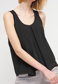 Černá bezramenní blůza s krajkovými akcenty na ramenech, s plisovaným předním designem, hladkým materiálem a volným střihem sahajícím pod pas.