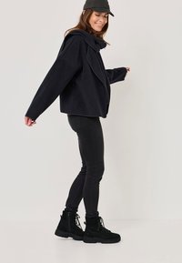 Giacca nera in misto lana con colletto oversize, abbinata a jeans neri slim-fit e stivaletti neri con lacci.