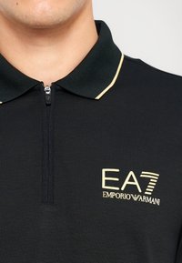 Juoda polo marškinėlių su užtrauktuku apykakle, dekoruoti auksinėmis detalėmis ir išryškinta "EA7 Emporio Armani" logotipu. Tekstūruota medžiaga.