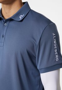 Mörkblå pikétröja i slätt tyg, med krage, tre knappar och broderad "J.LINDEBERG" på ärmen.