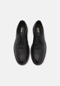 Une paire de chaussures habillées en cuir noir à lacets, avec des orteils ronds et la marque Clarks sur les semelles intérieures noires, vue de dessus.