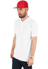 Valge puuvillane polo särk lühikeste varrukatega ja kahe nupuga, kantud tumedate teksade ja punase snapback mütsiga. Kuldsed kellad randmel.