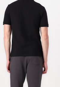 Homme portant un t-shirt noir uni à manches courtes et un pantalon gris foncé, debout de dos sur un fond blanc.