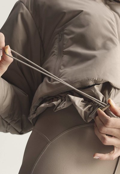 Person passt die Kordel an einer braunen gesteppte Jacke an, trägt passende braune Leggings, Nahaufnahme von Händen und Oberkörper.