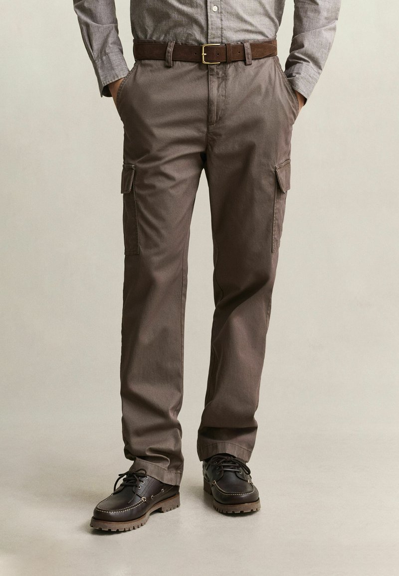 Hombre con pantalones cargo marrones, cinturón de cuero oscuro, camisa gris con las mangas remangadas y zapatos negros con cordones, de pie con las manos en los bolsillos.