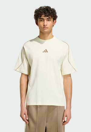 Ung mand med krøllet hår iført en cremefarvet Adidas T-shirt med brunt logo og matchende brune bukser, står mod en ensfarvet baggrund.