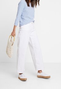 Pantalons larges blancs, haut rayé bleu et blanc à manches longues, baskets blanches avec semelle en espadrille, et un sac tissé beige.