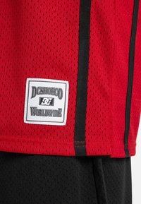 Červený športový top na ramienka s mriežkovou textúrou, s čiernymi bočnými akcentmi a bielou etiketou s textom "DC SHOE CO WORLDWIDE" na leme.