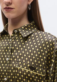 Brūna rakstaina krekls ar dzelteniem ģeometriskiem motīviem, standarta apkakli, pogu siju un kabatu ar kontrastējošām maliņām.