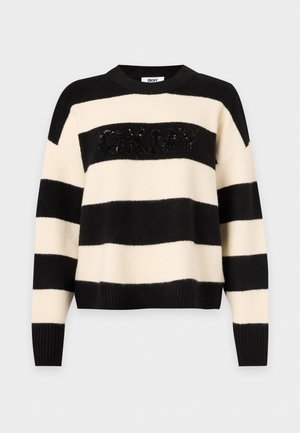 Femme portant un pull rayé noir et beige, un pantalon blanc à jambes larges, des chaussures noires pointues, tenant un sac à main noir d'une main.
