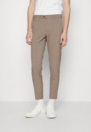 Homme portant un pantalon marron slim, des chaussettes blanches, des baskets blanches et un t-shirt blanc sans manches, debout devant un fond uni.