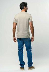 Bežs īsās piedurknes krekls ar taisnu griezumu, savienots ar zilajiem džinsiem un gaiši brūnām kurpēm, attēlots no aizmugures.