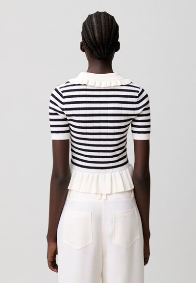 Mujer con el cabello trenzado que lleva una blusa de manga corta con rayas blancas y negras, con cuello y bajo con volantes, combinada con pantalones blancos de tiro alto.