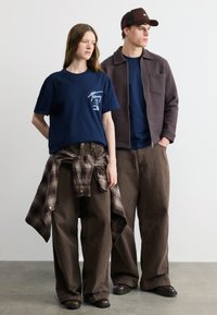 Δύο μοντέλα στέκονται δίπλα δίπλα, φορόντας καφέ παντελόνες με φαρδιά πόδια και μπλε μπλούζες· η γυναίκα με πουκάμισο καρό δεμένο γύρω από τη μέση, ο άντρας με καφέ τζάκετ και καπέλο.