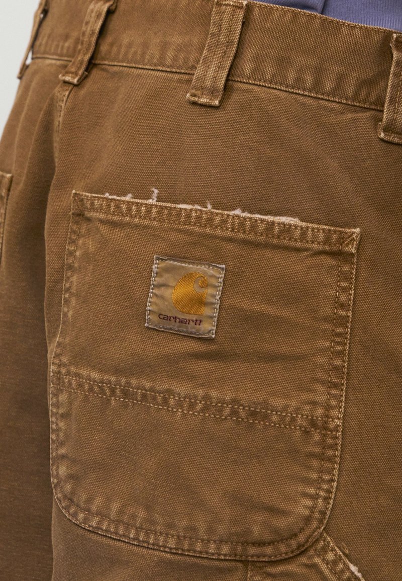 Primer plano del bolsillo trasero de unos pantalones marrones Carhartt con etiqueta de marca visible y detalle de borde deshilachado.