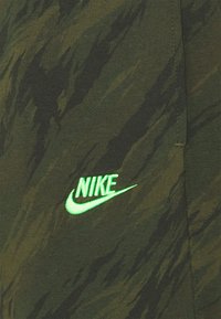Zelená látka s maskáčovým vzorem, která má na levé straně zářící zelené logo Nike, s jemnou texturou a volným střihem.