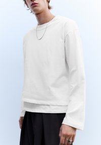 Weißes Langarm-T-Shirt mit entspannter Passform, rundem Ausschnitt und leicht strukturierter Stoffqualität, kombiniert mit dunklen, locker sitzenden Hosen.