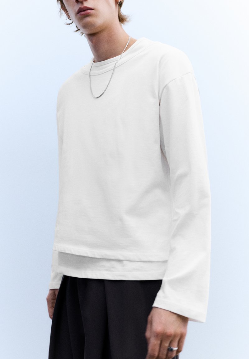 Weißes Langarm-T-Shirt mit entspannter Passform, rundem Ausschnitt und leicht strukturierter Stoffqualität, kombiniert mit dunklen, locker sitzenden Hosen.