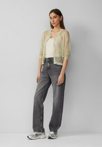 Lichtgroene, gebreide cardigan met knopen, gedragen over een crèmekleurig tanktop. Draagt het met losse grijze jeans en metallic sneakers.